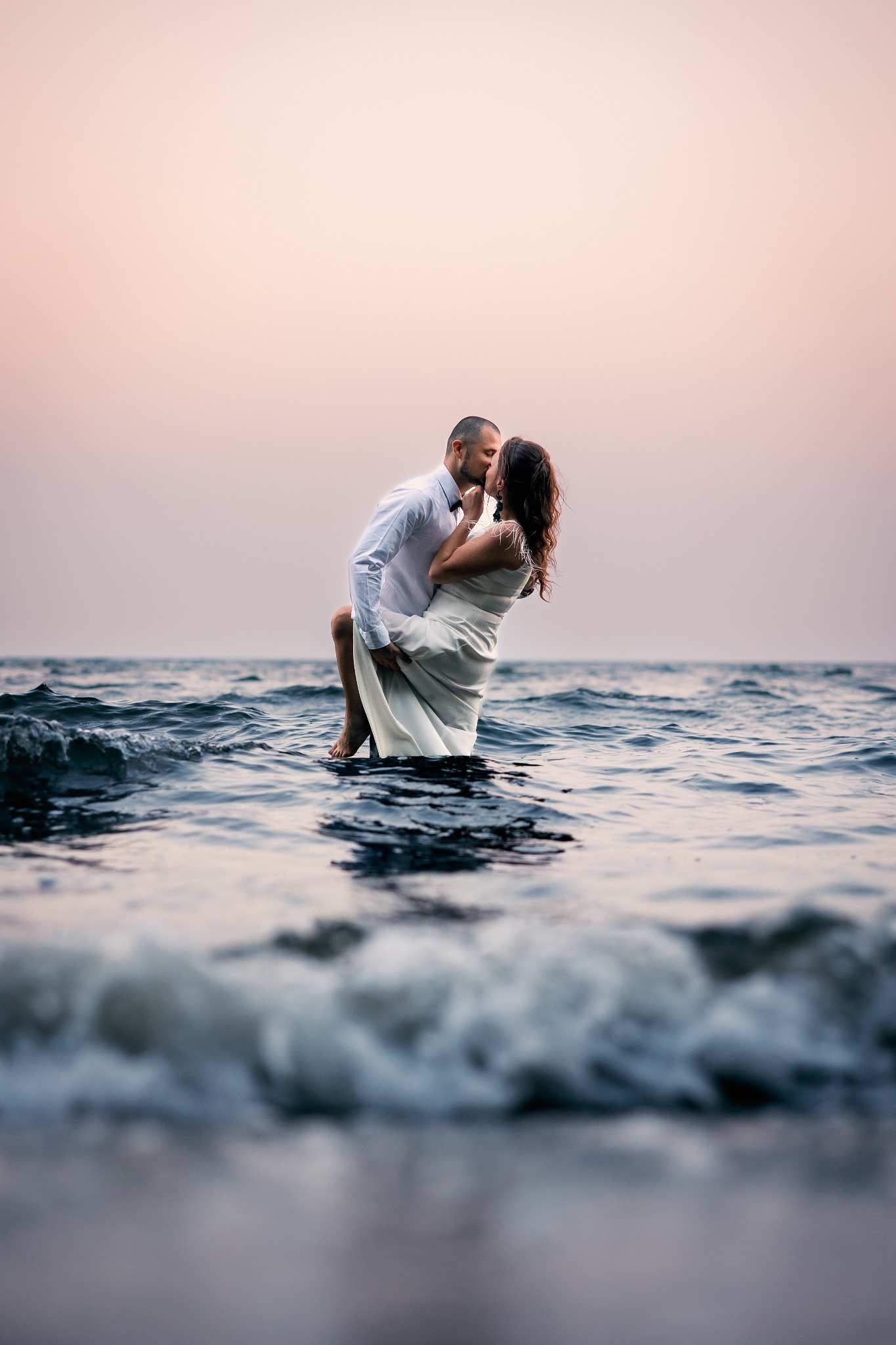 Para młoda w objęciach, pocałunek w wodzie o zmierzchu. Mężczyzna w białej koszuli trzyma kobietę w białej sukni. Romantyczna sesja na tle spokojnego morza.