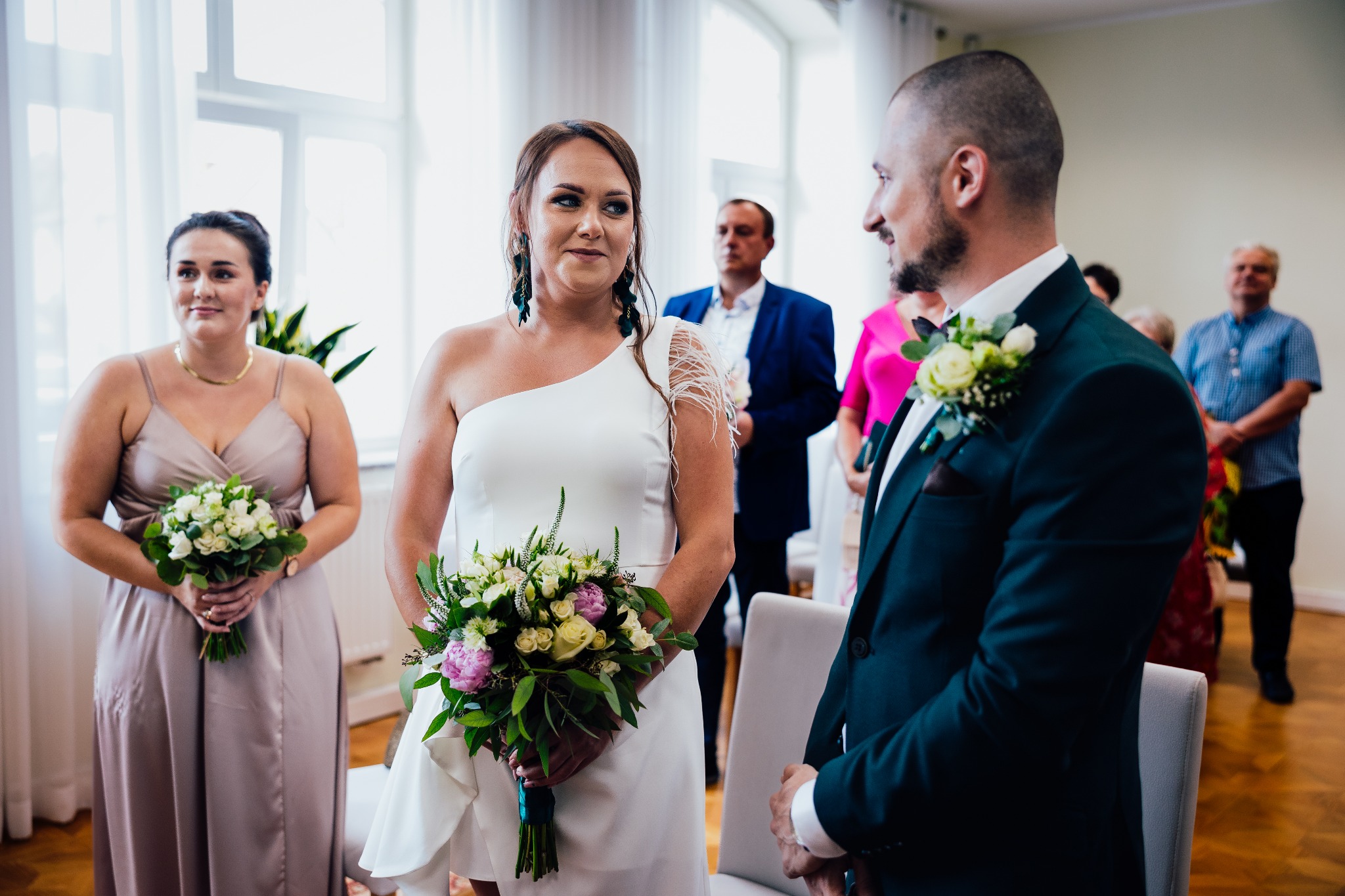 Para młoda podczas ceremonii ślubnej w jasnym wnętrzu, panna młoda w białej sukni z bukietem, pan młody w ciemnozielonym garniturze z butonierką. Goście w tle.