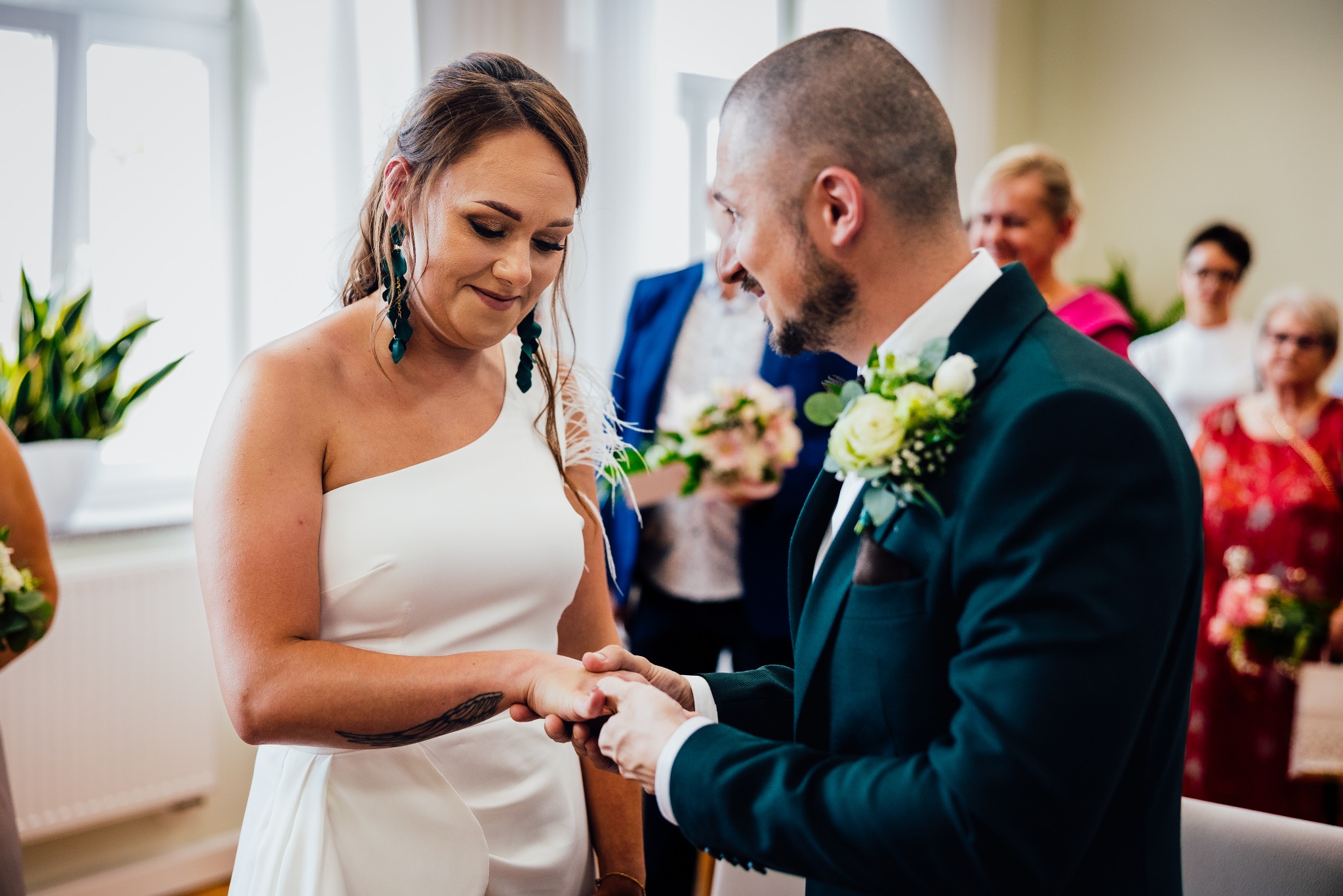 Panna młoda w białej sukni z odkrytym ramieniem i tatuażem na ramieniu, podczas ceremonii ślubnej, patrzy na dłoń, na którą pan młody zakłada obrączkę.