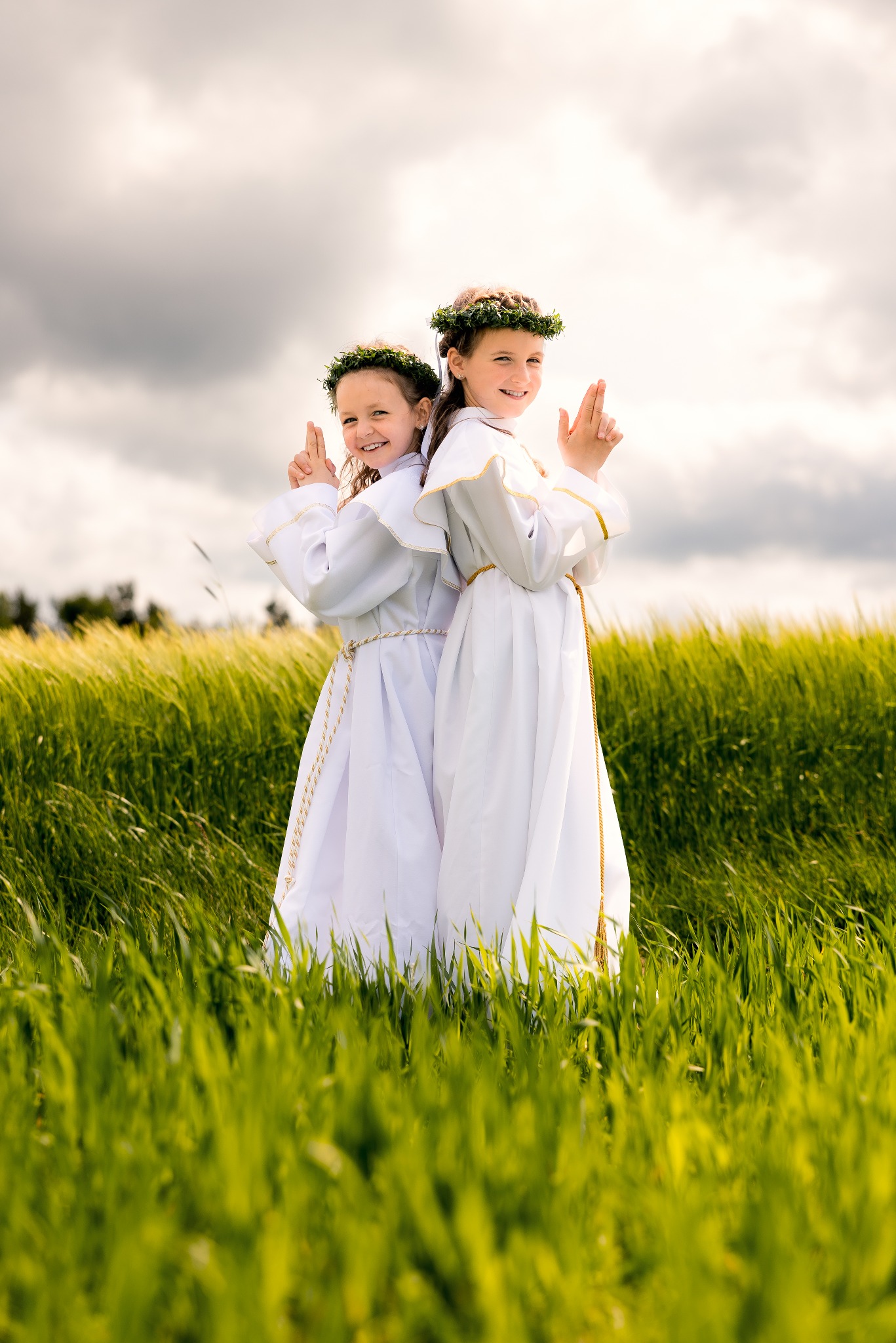 Dwie uśmiechnięte dziewczynki w białych strojach komunijnych i wiankach na głowach, stojące tyłem do siebie na tle zielonego pola i pochmurnego nieba.