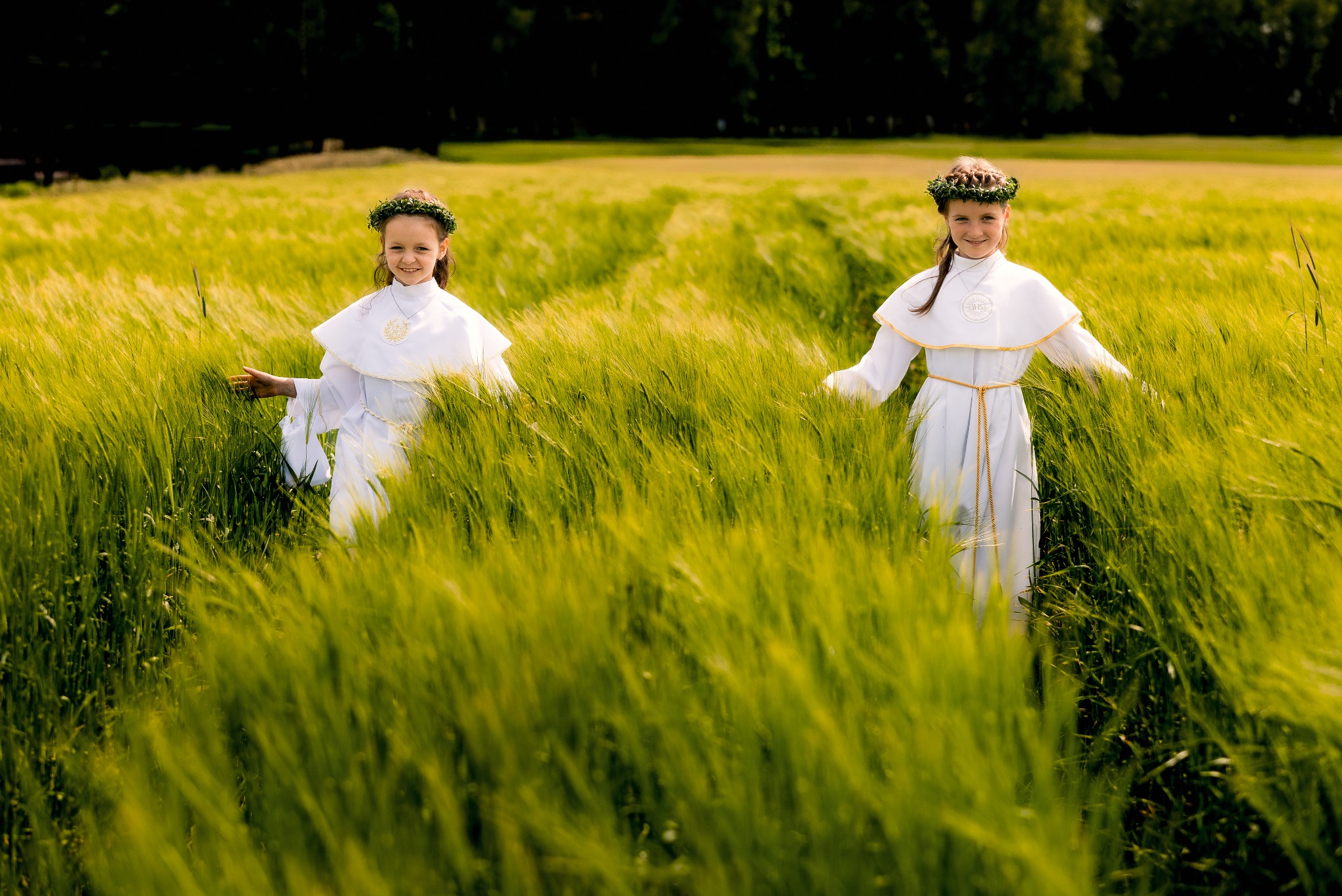 Dwie dziewczynki w białych strojach komunijnych i wiankach na głowach stoją na zielonym polu zboża w słoneczny dzień. Uśmiechnięte, z rozłożonymi rękami.