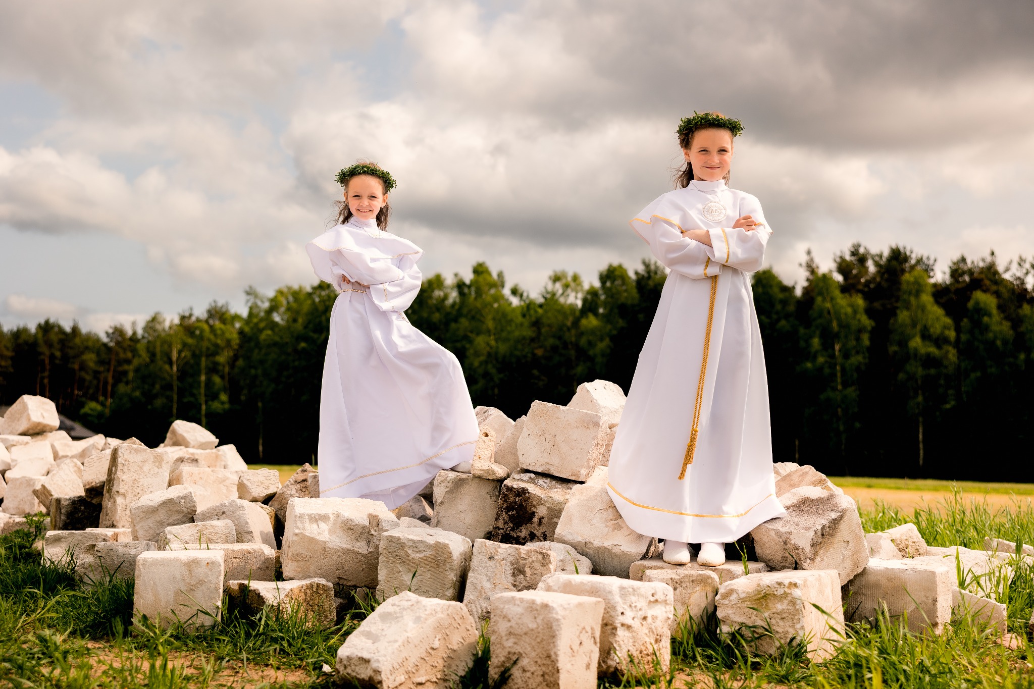 Dwie dziewczynki w białych strojach komunijnych, pozujące na tle sterty kamieni i zieleni. Uśmiechnięte, z wiankami na głowach, na tle pochmurnego nieba. Sesja komunijna w plenerze.