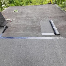 Płaski dach pokryty papą termozgrzewalną, częściowo zabezpieczony taśmą uszczelniającą; widoczne narzędzia, rękawice robocze i rolka papy na tle zieleni drzew.