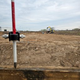 GEOSTREFA Usługi Geodezyjne - Prace geodezyjne na działce budowlanej. W tle minikoparka i pracownik. W pierwszym planie tyczka geodezyjna z bolcem wbita w drewnianą deskę.