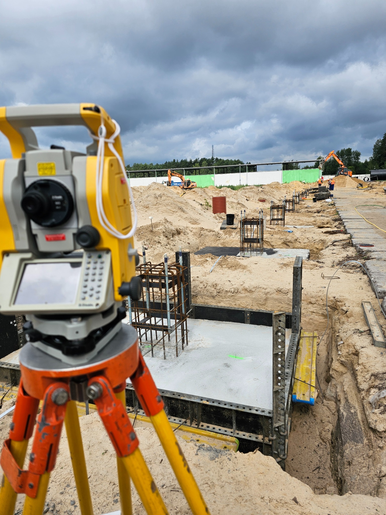 Żółty teodolit na trójnogu, w tle wykopy pod fundamenty zbrojone stalą, koparka i pochmurne niebo. Prace ziemne na budowie.
