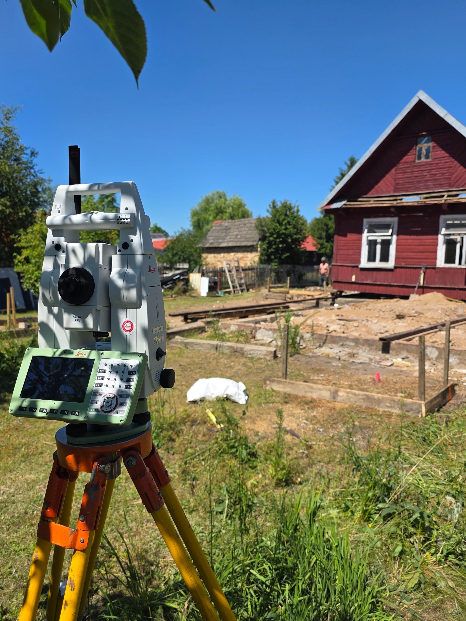 Tachimetr Leica na statywie, w tle czerwony dom w trakcie renowacji fundamentów. Widoczne drewniane konstrukcje i osoba w oddali. Słoneczny dzień.