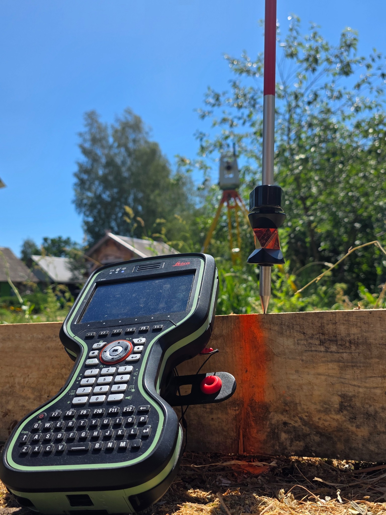 Pomiar geodezyjny: tachimetr, tyczka z pryzmatem i kontroler Leica na tle drewnianej konstrukcji i zieleni. Pomiar wytyczenia budynku w terenie.