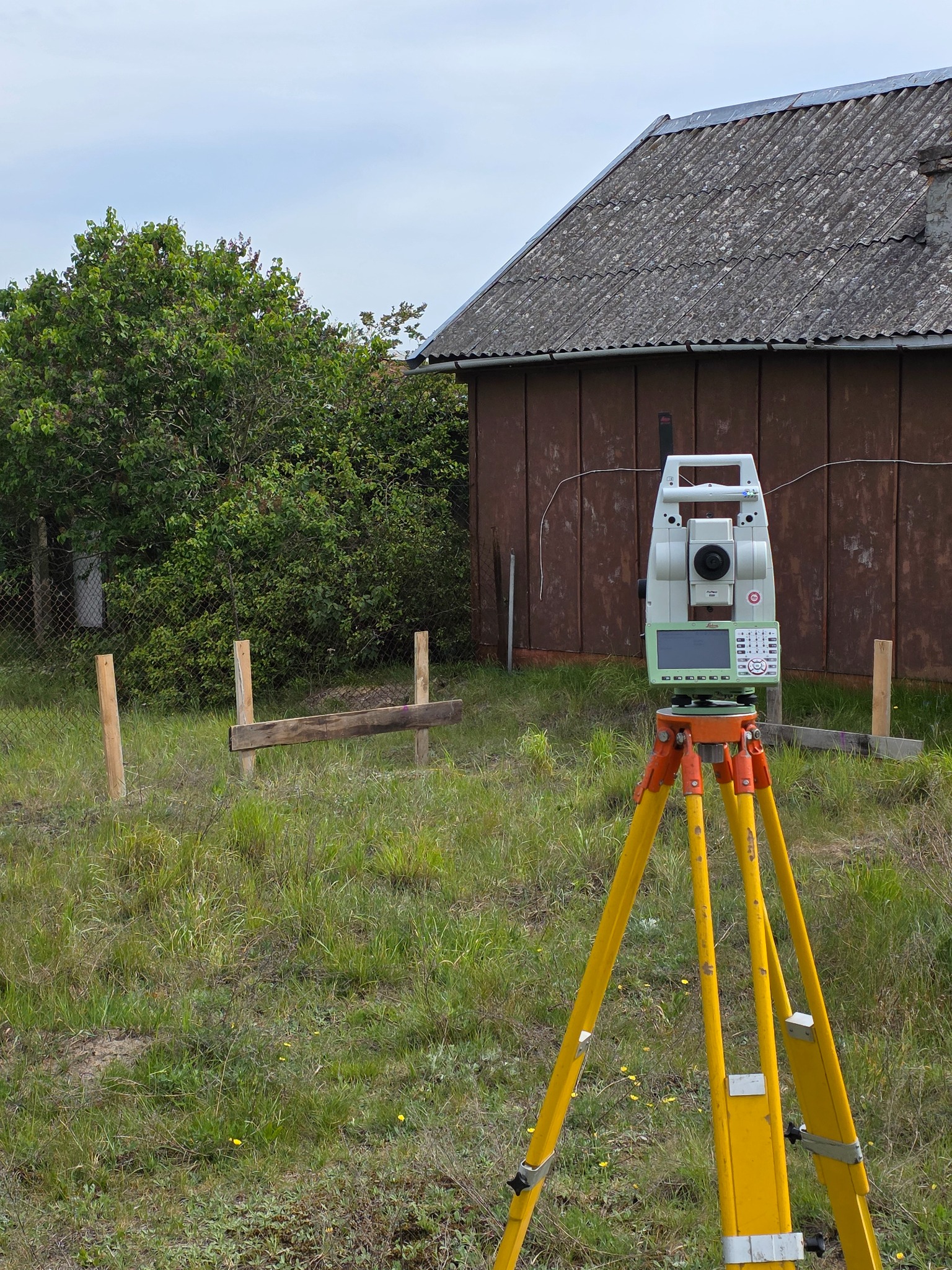 Żółty teodolit na trójnogu ustawiony na łące, w tle drewniany budynek i zielone drzewa. Widoczne drewniane słupki wyznaczające teren.