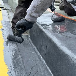 Kempol Sp. Z o.o. - Aplikacja szarej hydroizolacji na dachu płaskim za pomocą wałka, widoczne detale wykończeniowe i żółta linia bezpieczeństwa.