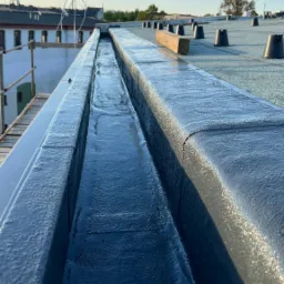 Widok z góry na pomalowany na czarno kanał odpływowy na dachu budynku, z widocznymi elementami mocującymi i drewnianą belką, w tle budynki i błękitne niebo.