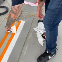 Pracownik w jeansach aplikuje masę wyrównującą na betonowej powierzchni tarasu za pomocą szpachelki, widoczne taśmy malarskie, rękawice ochronne i odkurzacz przemysłowy w tle.