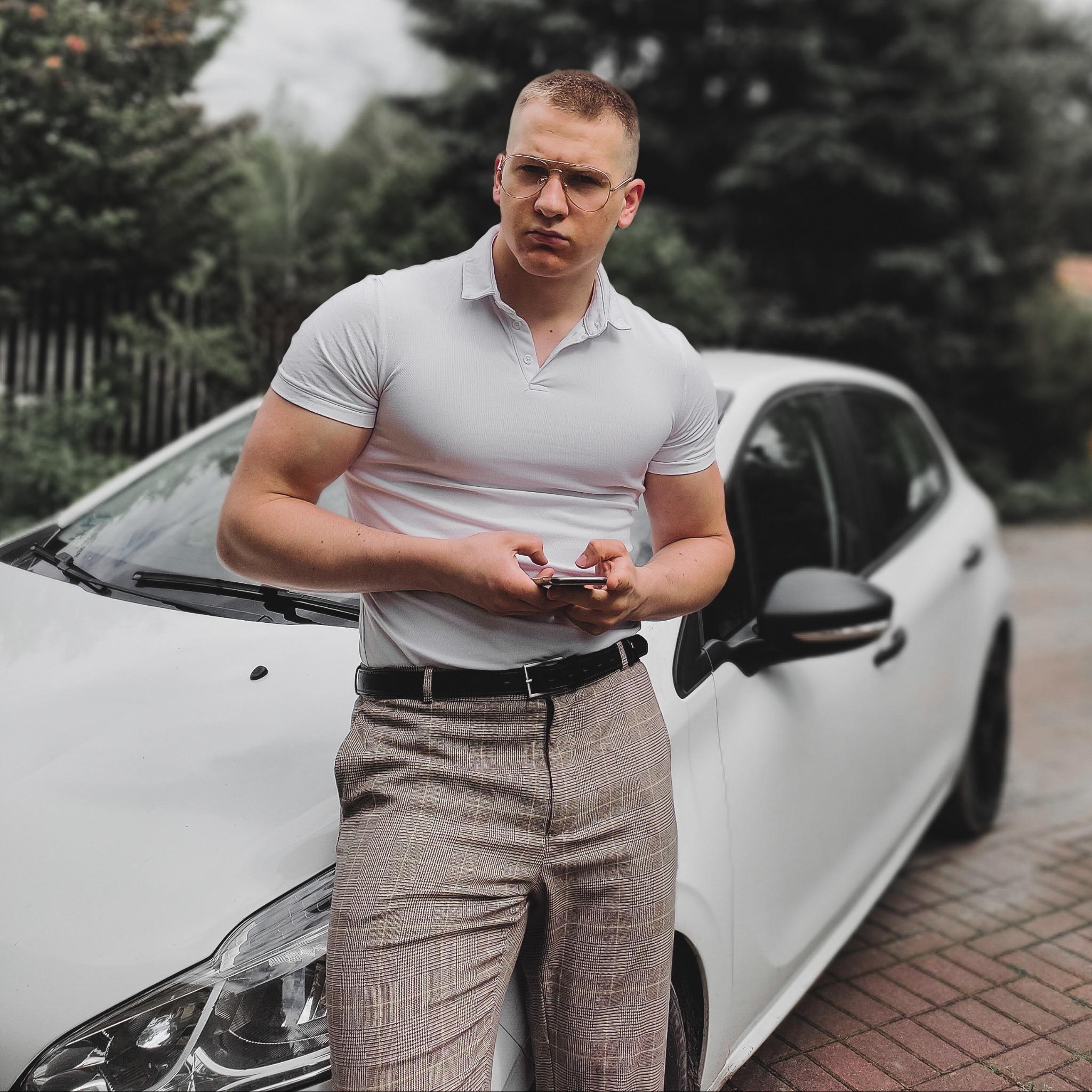 Mężczyzna w okularach i białej koszulce polo opiera się o biały samochód, patrząc w bok i trzymając telefon komórkowy.