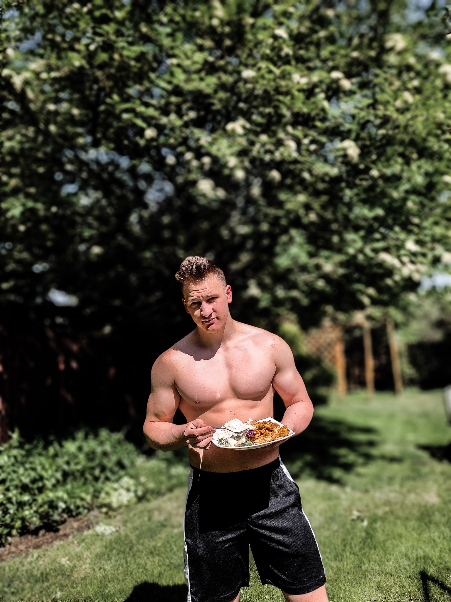 Mężczyzna o muskularnej sylwetce, trzymający talerz z posiłkiem na trawie, w tle drzewo.