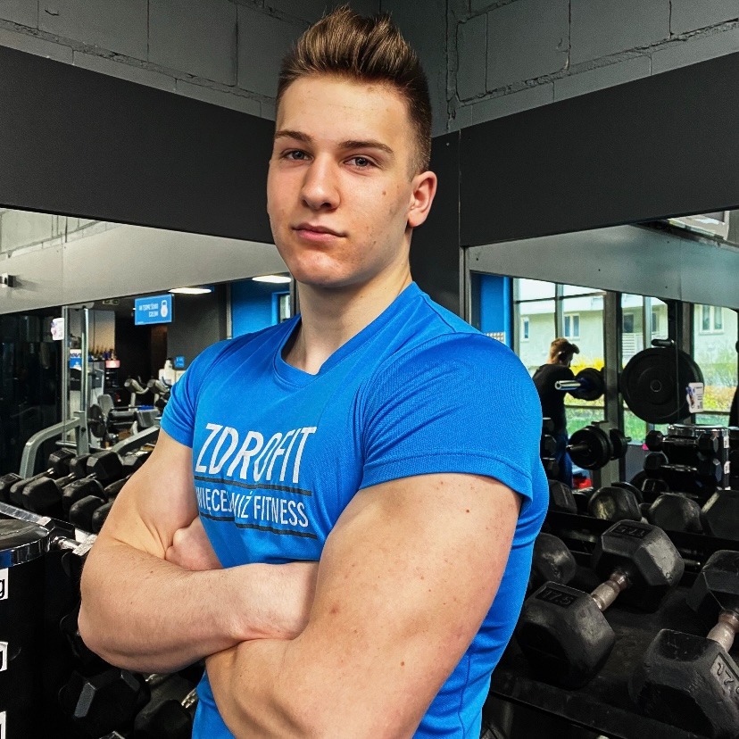 Młody mężczyzna z założonymi rękami w niebieskiej koszulce z nadrukiem, prezentujący sylwetkę w tle siłowni z hantlami i lustrami.