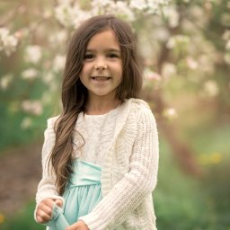 Sesje ciążowe Rzeszów - Monika Paradysz - Uśmiechnięta dziewczynka w białej sukience z błękitnym pasem i kremowym sweterku, pozująca na tle kwitnącego drzewa.