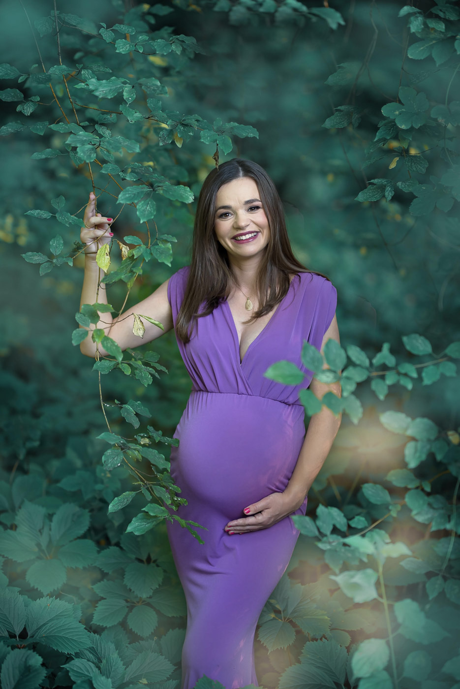 Uśmiechnięta kobieta w liliowej sukni ciążowej, pozująca w plenerze, otoczona zielenią liści i gałęzi.