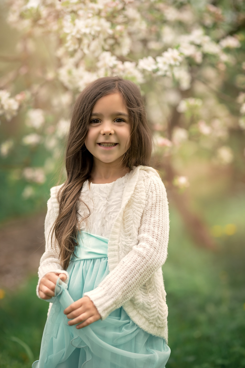 Uśmiechnięta dziewczynka w białej sukience z błękitnym pasem i kremowym sweterku, pozująca na tle kwitnącego drzewa.
