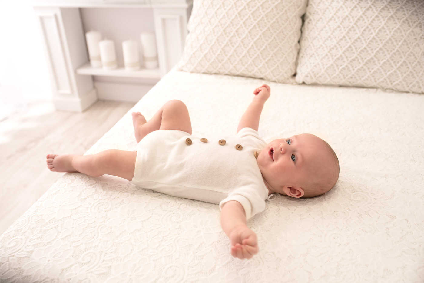 Niemowlę leżące na plecach na białej, koronkowej narzucie, ubrane w białe body z drewnianymi guzikami, z uniesionymi rączkami i uśmiechem na twarzy, w tle białe meble.