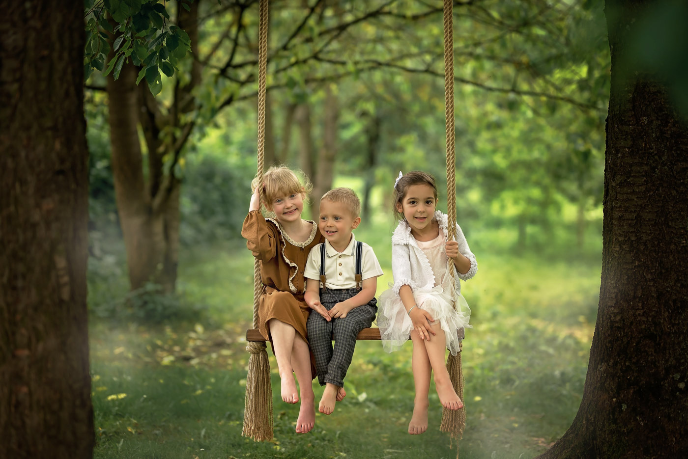 Troje uśmiechniętych dzieci siedzi na drewnianej huśtawce zawieszonej na linach między drzewami w plenerze.