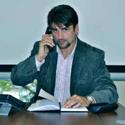 Mężczyzna w sztruksowej marynarce rozmawia przez telefon, notując w otwartym notatniku na biurku, z widocznym zegarkiem na lewym nadgarstku.