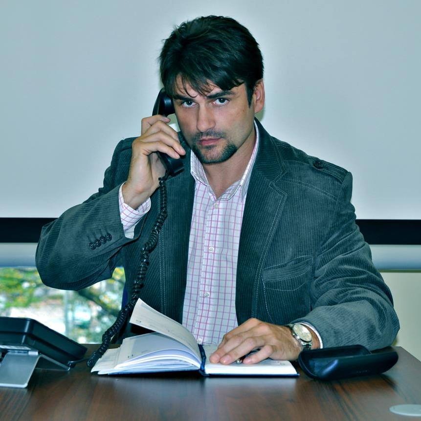 Mężczyzna w sztruksowej marynarce rozmawia przez telefon, notując w otwartym notatniku na biurku, z widocznym zegarkiem na lewym nadgarstku.