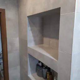 Szara łazienka z wnęką wyłożoną płytkami, na półkach kosmetyki i akcesoria do pielęgnacji.