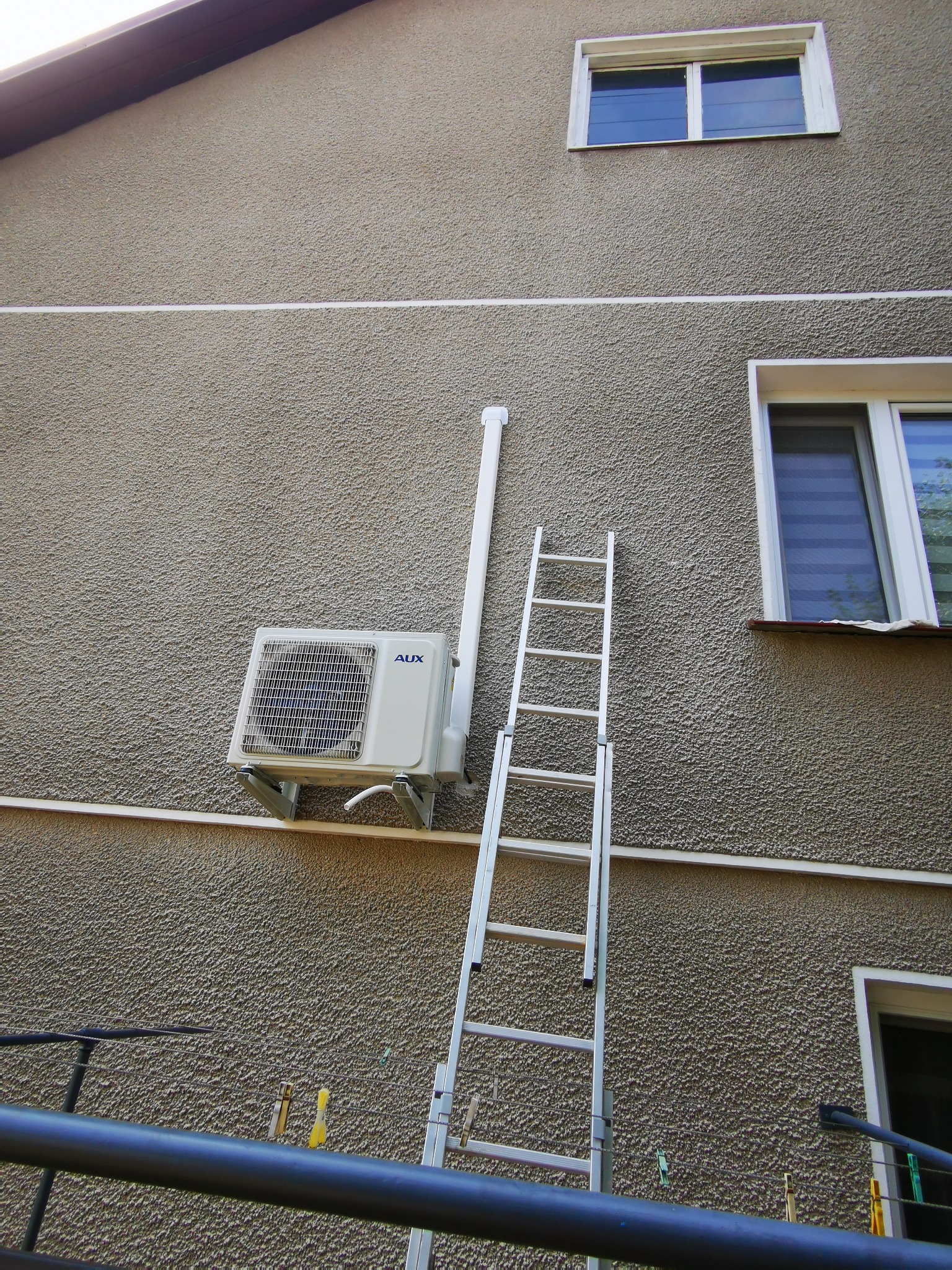 Zamontowana jednostka zewnętrzna klimatyzatora marki AUX na teksturowanej elewacji budynku, z drabina opartą o ścianę i widoczną rurą odprowadzającą skropliny.