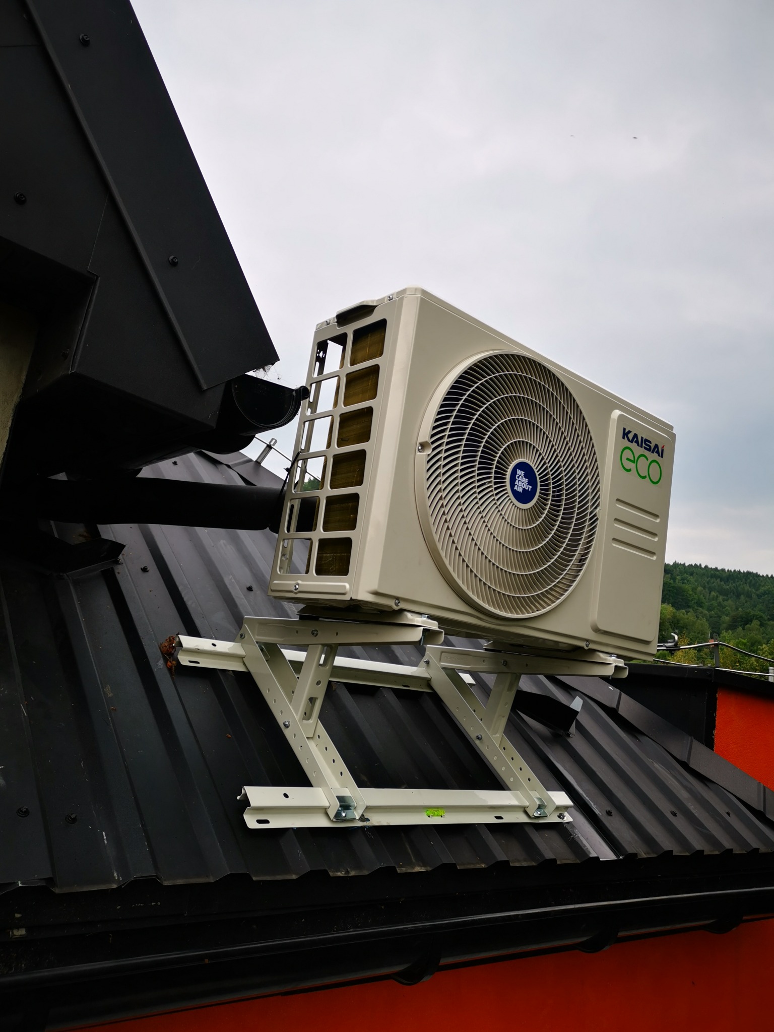 Jednostka zewnętrzna klimatyzatora Kaisai ECO zamontowana na dachu budynku na specjalnym, białym stelażu. Widoczny czarny dach z blachy trapezowej i fragment czerwonej ściany budynku.