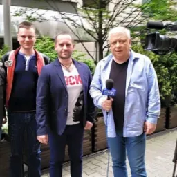 Czterech mężczyzn pozuje do zdjęcia przed kamerą telewizyjną z mikrofonem TVP, na tle zieleni.