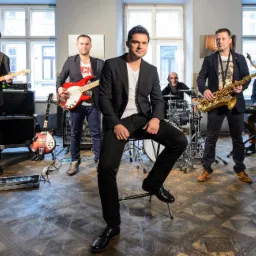 Zespół muzyczny w składzie: gitarzysta z gitarą elektryczną, basista z gitarą basową, perkusista za perkusją, saksofonista z saksofonem i klawiszowiec z keyboardem, lider zespołu siedzący...
