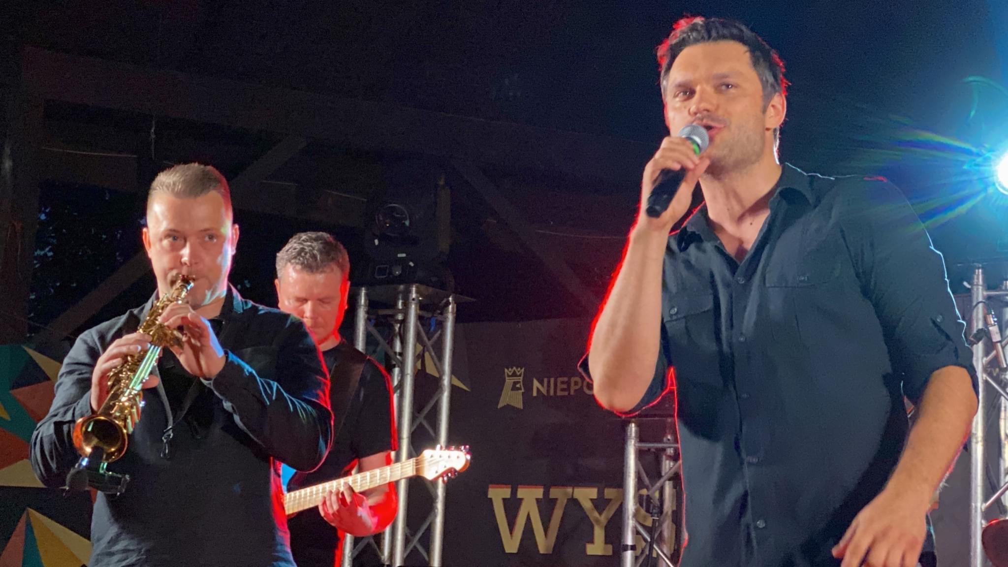 Zespół muzyczny na scenie: wokalista z mikrofonem, saksofonista i gitarzysta, oświetlenie sceniczne z tyłu.