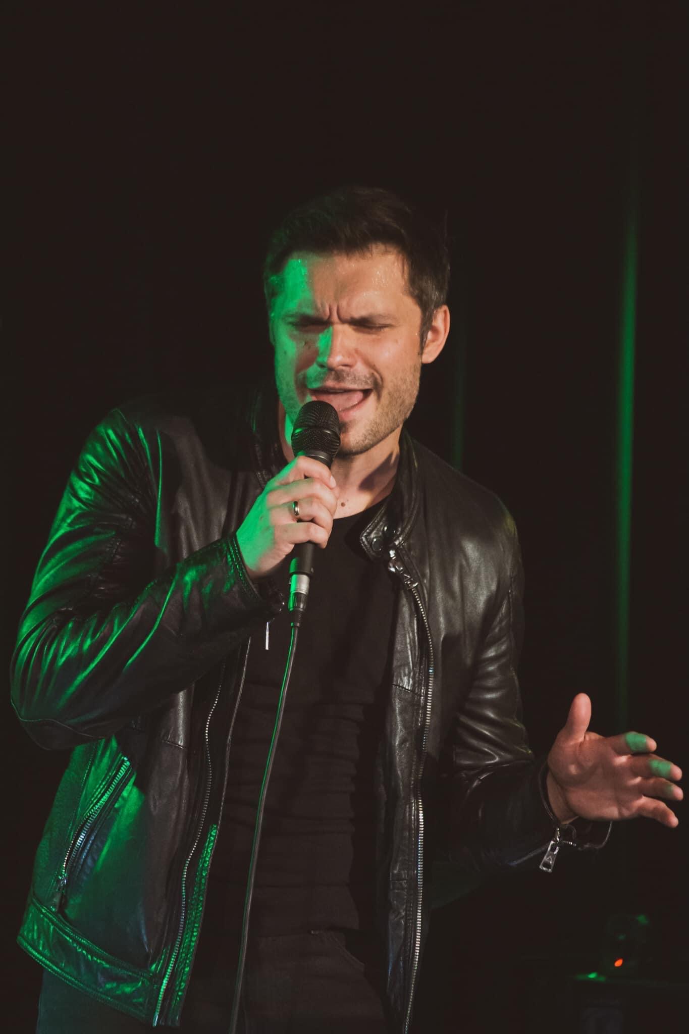 Wokalista w czarnej skórzanej kurtce śpiewa do mikrofonu, oświetlony zielonym światłem scenicznym, z widocznym pierścieniem na palcu.