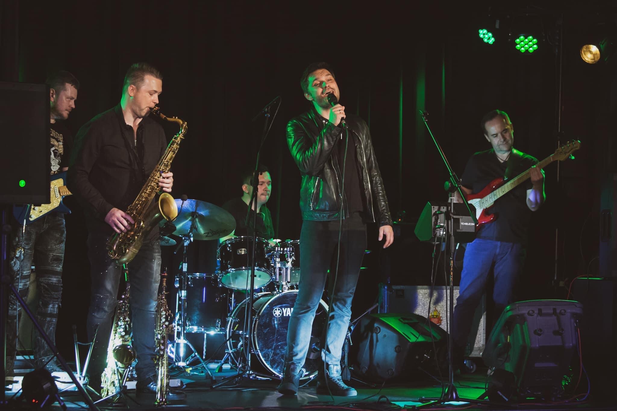 Zespół muzyczny na scenie podczas występu: wokalista w skórzanej kurtce, saksofonista, gitarzysta i perkusista, oświetlenie sceniczne w odcieniach zieleni.