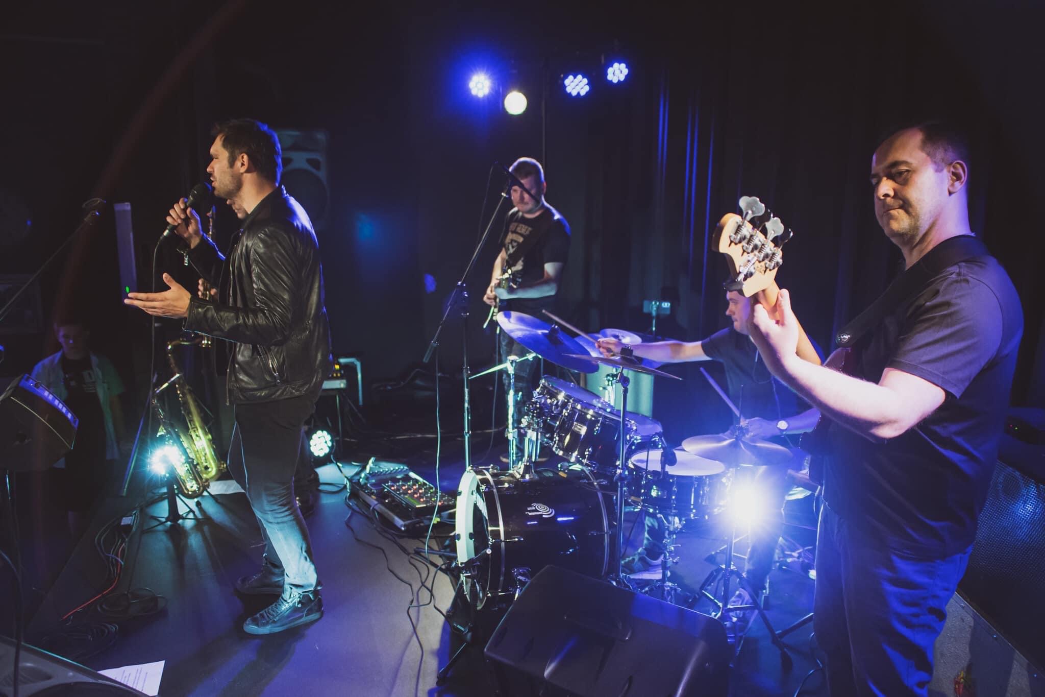 Zespół muzyczny na scenie podczas występu, wokalista w skórzanej kurtce gestykuluje, w tle perkusista i gitarzysta, oświetlenie sceniczne w odcieniach niebieskiego.