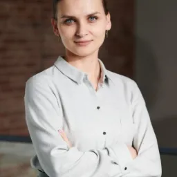 Portret kobiety w jasnej koszuli z założonymi rękami na tle ceglanej ściany i szarego tła.