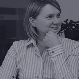 Kobieta w bluzce w paski z długopisem i otwartą książką, oparta podbródkiem na dłoni, spogląda w bok z lekkim uśmiechem.