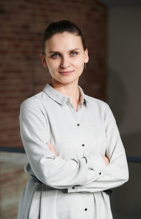 Portret kobiety w jasnej koszuli z założonymi rękami na tle ceglanej ściany i szarego tła.