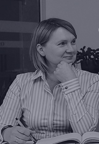 Kobieta w bluzce w paski z długopisem i otwartą książką, oparta podbródkiem na dłoni, spogląda w bok z lekkim uśmiechem.