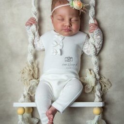 Anna Wańczyk Fotografia - Śpiąca noworodka w białym ubranku z koronkowymi rękawami i kwiatową opaską na głowie, pozuje na mini-huśtawce z makramy na neutralnym tle.