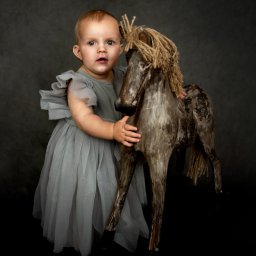 Anna Wańczyk Fotografia - Urocza dziewczynka w tiulowej sukience przytula stylizowanego, drewnianego konika na biegunach z jutową grzywą, na ciemnym tle.