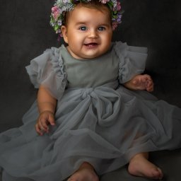 Anna Wańczyk Fotografia - Uśmiechnięta niemowlęca dziewczynka w szarej tiulowej sukience i wianku z kwiatów na głowie, siedząca na szarym tle.