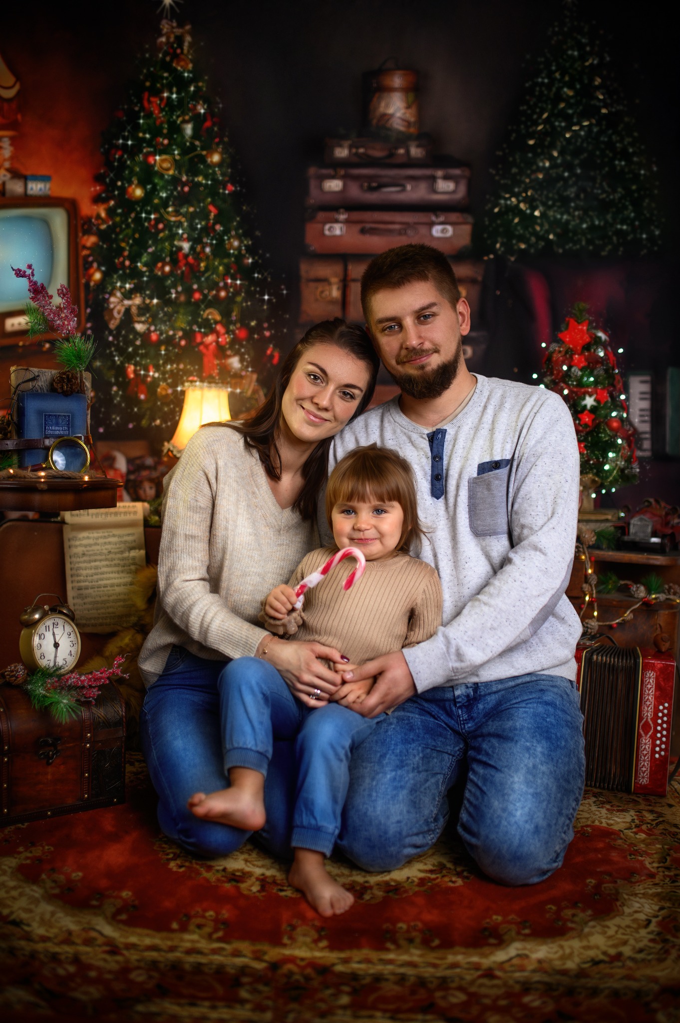 Rodzina, mama, tata i córka, pozują w świątecznej scenerii z choinkami, starym telewizorem i walizkami w tle; dziewczynka trzyma lizaka.