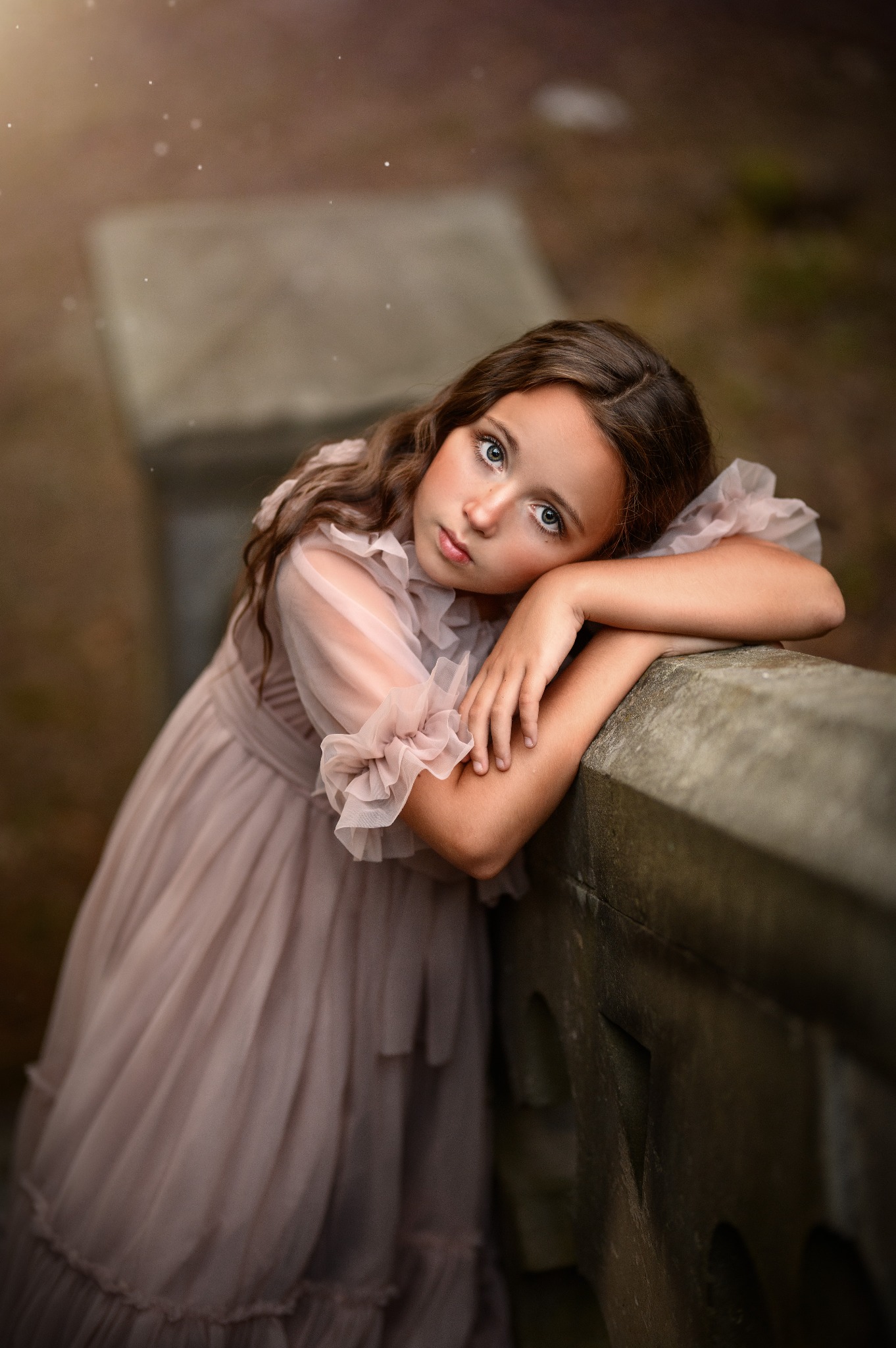 Portret dziewczynki w zwiewnej, pudroworóżowej sukience z falbanami, opierającej się o kamienną balustradę, z zamyślonym spojrzeniem i delikatnym makijażem.