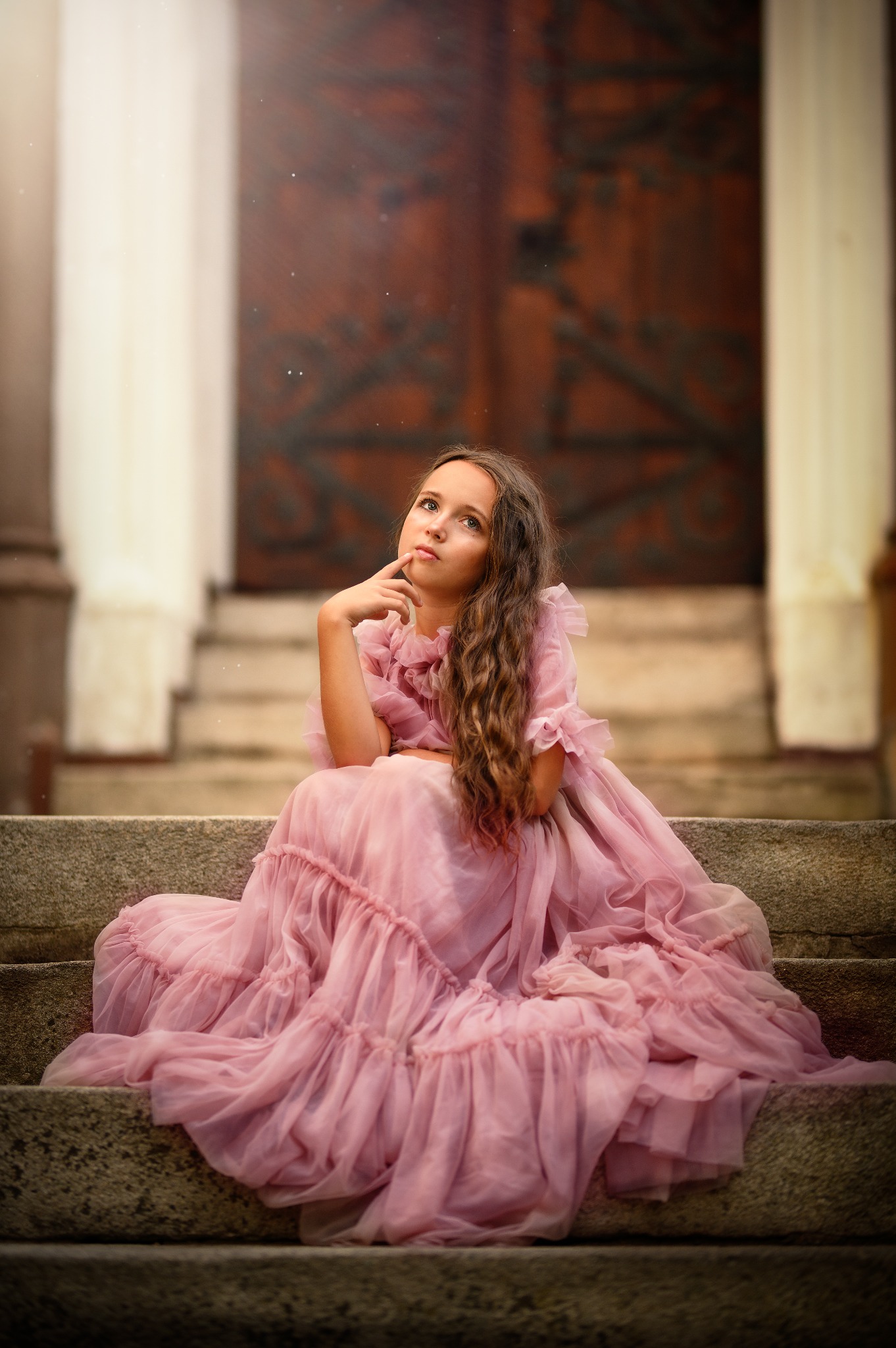 Młoda dziewczyna w długiej, różowej sukni siedzi na kamiennych schodach, zamyślona, z palcem wskazującym na ustach, na tle drewnianych drzwi.