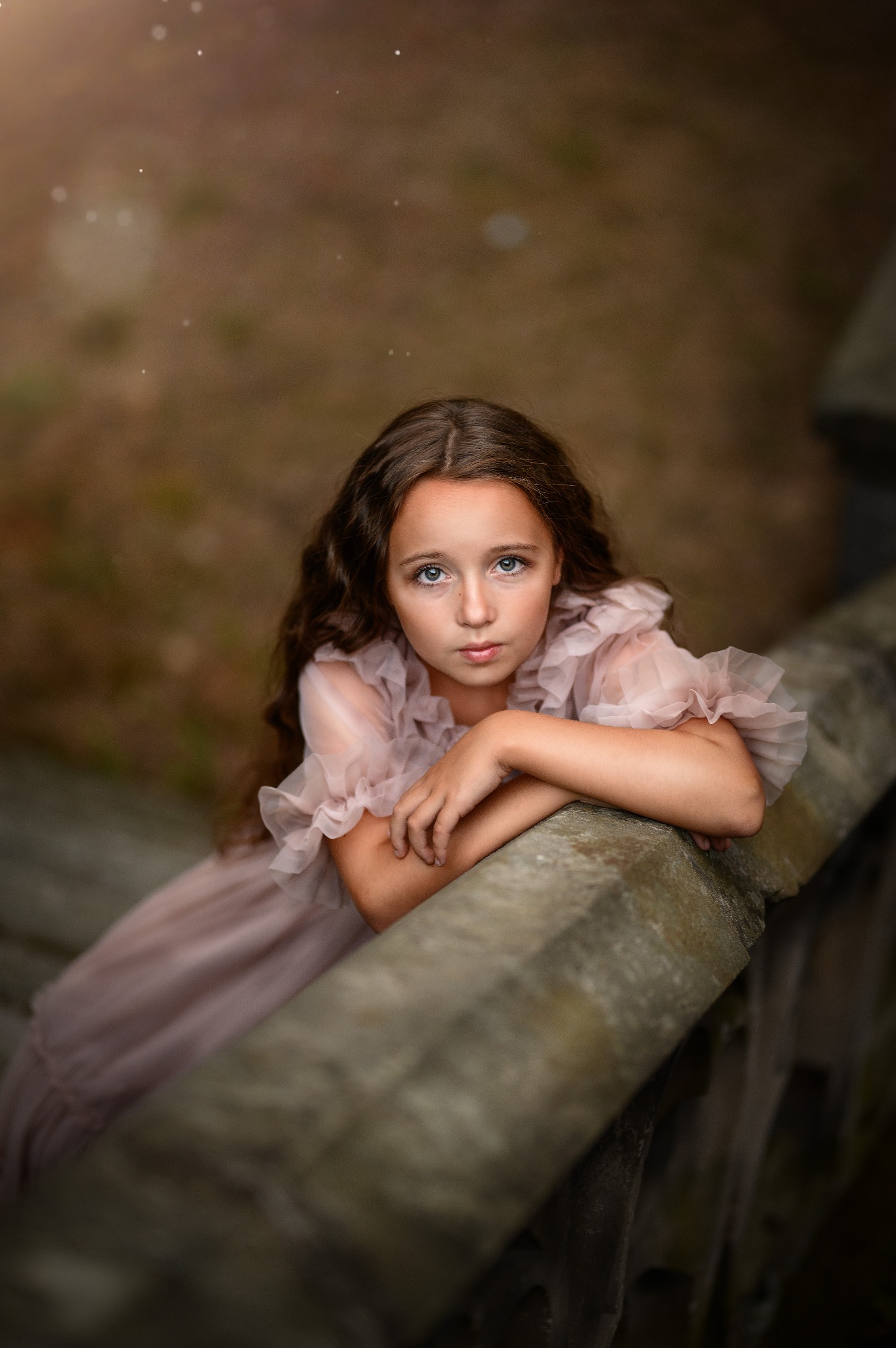 Portret młodej dziewczyny w zwiewnej, pudrowej sukience, opierającej się o kamienną balustradę, z głębokim spojrzeniem błękitnych oczu skierowanym w górę.