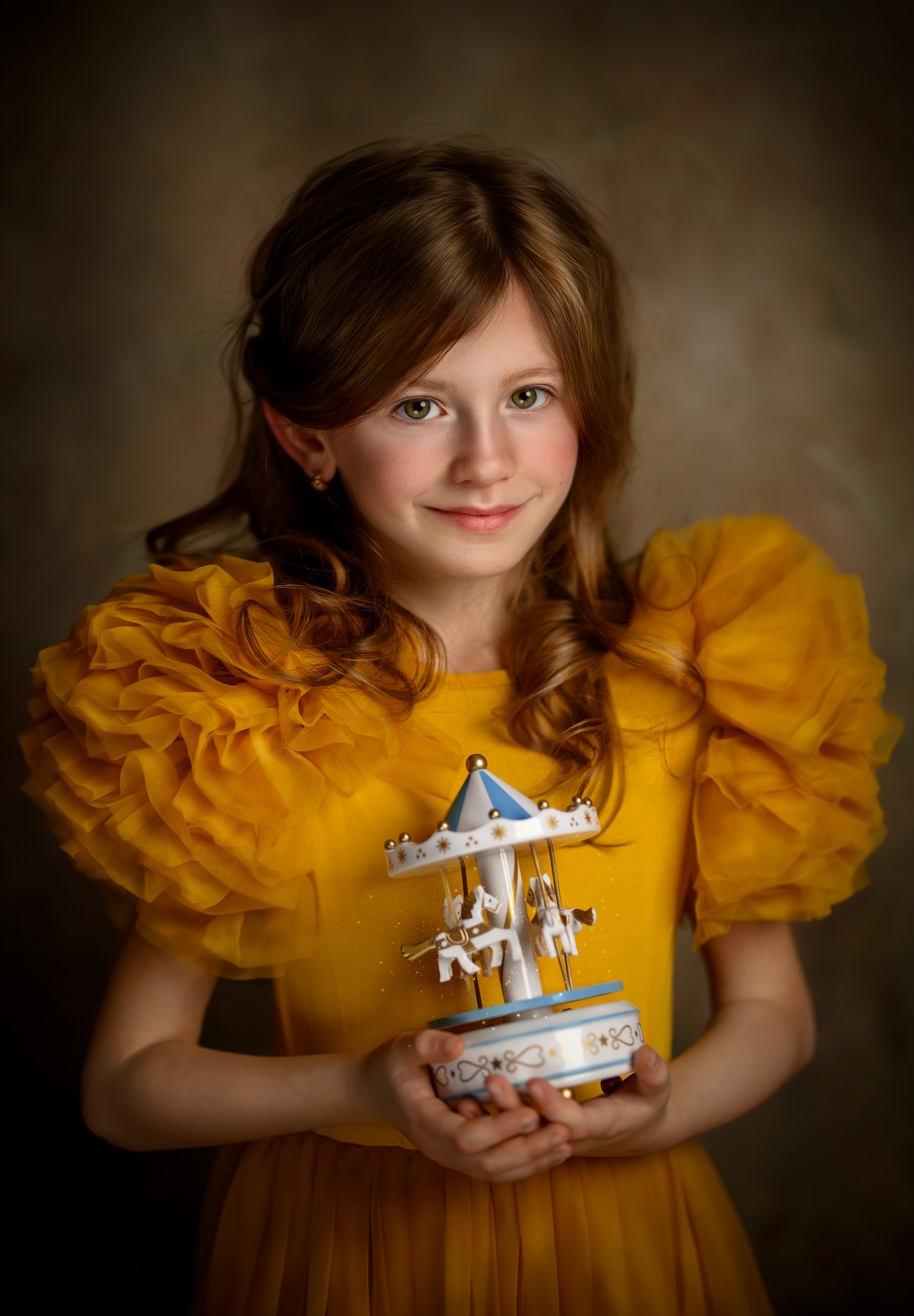 Portret uśmiechniętej dziewczynki w żółtej sukience z falbanami, trzymającej w dłoniach małą, ozdobną karuzelę z konikami, na rozmytym, ciemnym tle.
