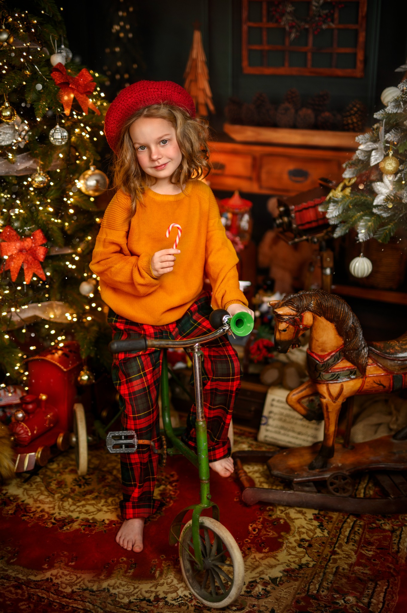 Dziewczynka w czerwonym berecie, żółtym swetrze i spodniach w kratę siedzi na zielonym rowerku z lizakiem na tle świątecznych dekoracji i choinek.