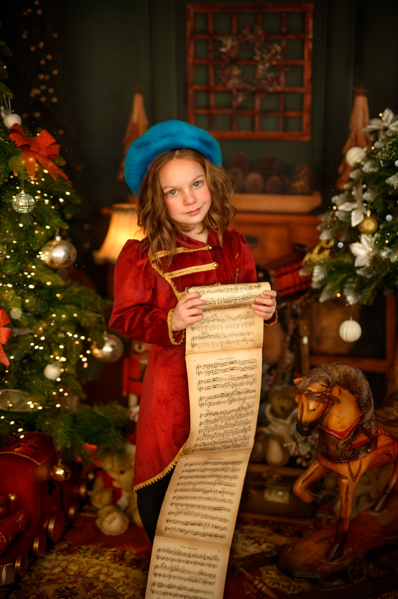 Dziewczynka w czerwonym mundurku i błękitnym berecie trzyma zwój nut na tle świątecznej dekoracji z choinkami i konikiem na biegunach.