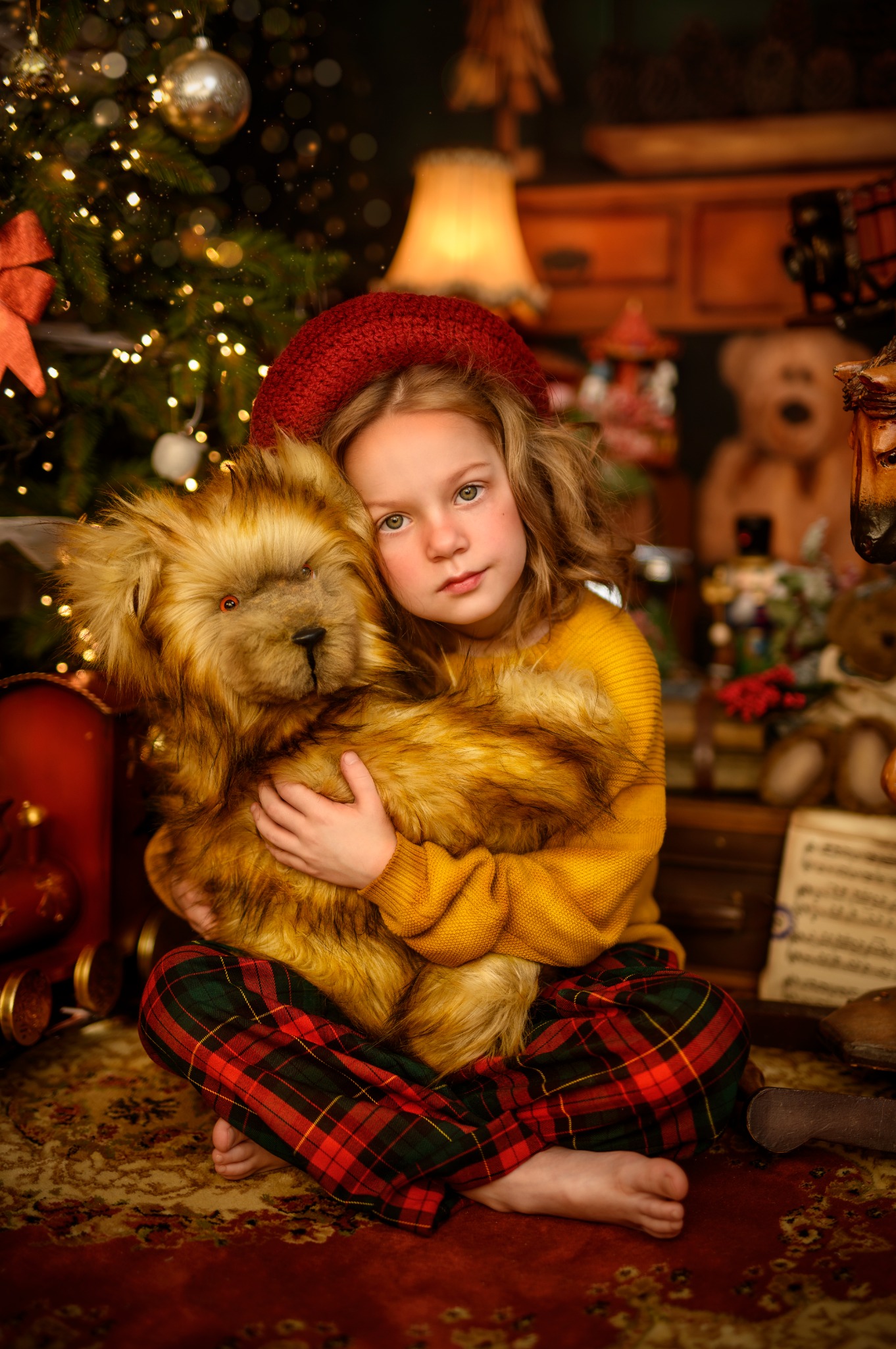 Dziewczynka w czerwonym berecie i żółtym swetrze, siedząca na podłodze w kratę, obejmująca pluszowego misia na tle świątecznej choinki i drewnianych mebli.