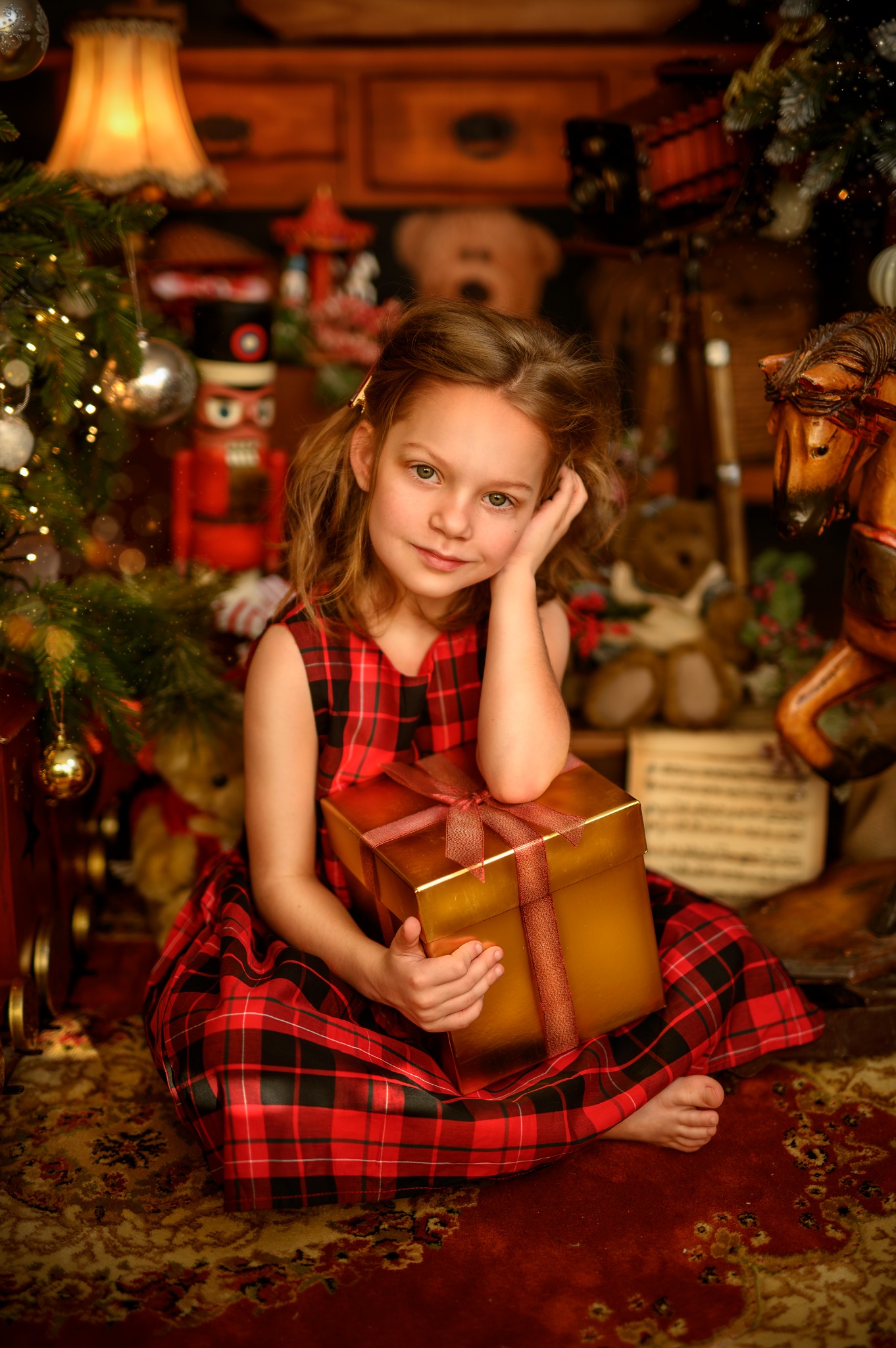 Dziewczynka w czerwonej sukience w kratę siedzi na dywanie z prezentem w złotym pudełku, obok choinki i drewnianego konika na biegunach, w tle zabawki i lampka.